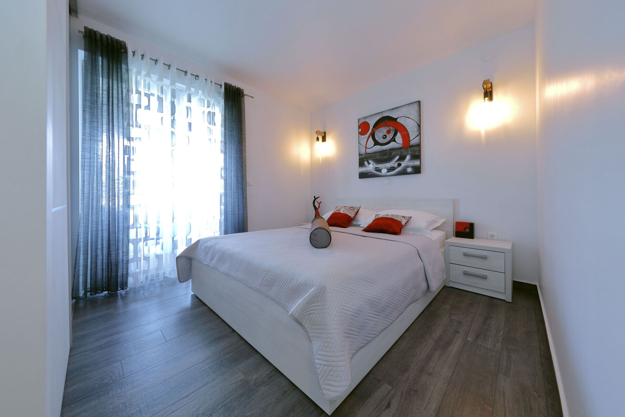 A modern bedroom with a large bed, a nightstand, a wall-mounted lamp, and a window with curtains.