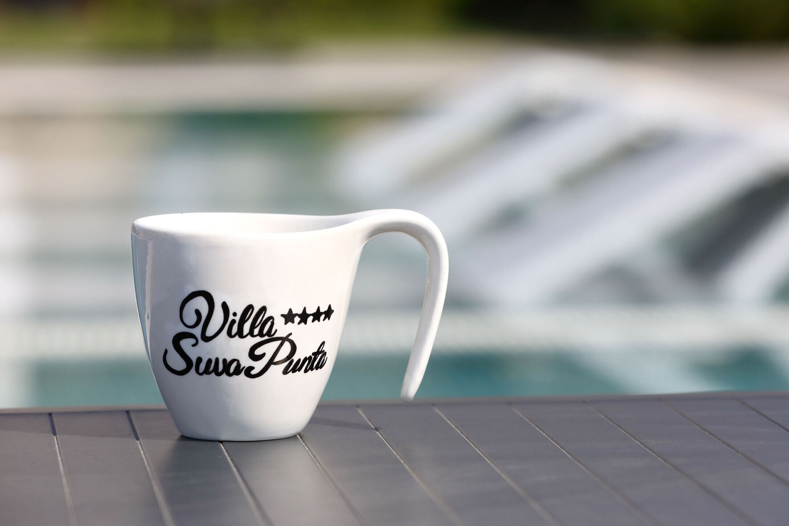 A white mug with 'Villa Swa Punta' text on a table near a pool.