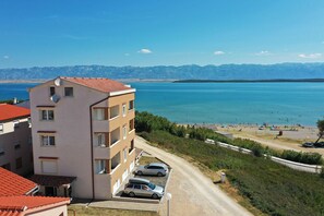 A multi-story building with a red-tiled roof, a parking area with cars, and a view of a lake and mountains in the background. - Apartment Sandstrand 2 with Sea View (Nin)