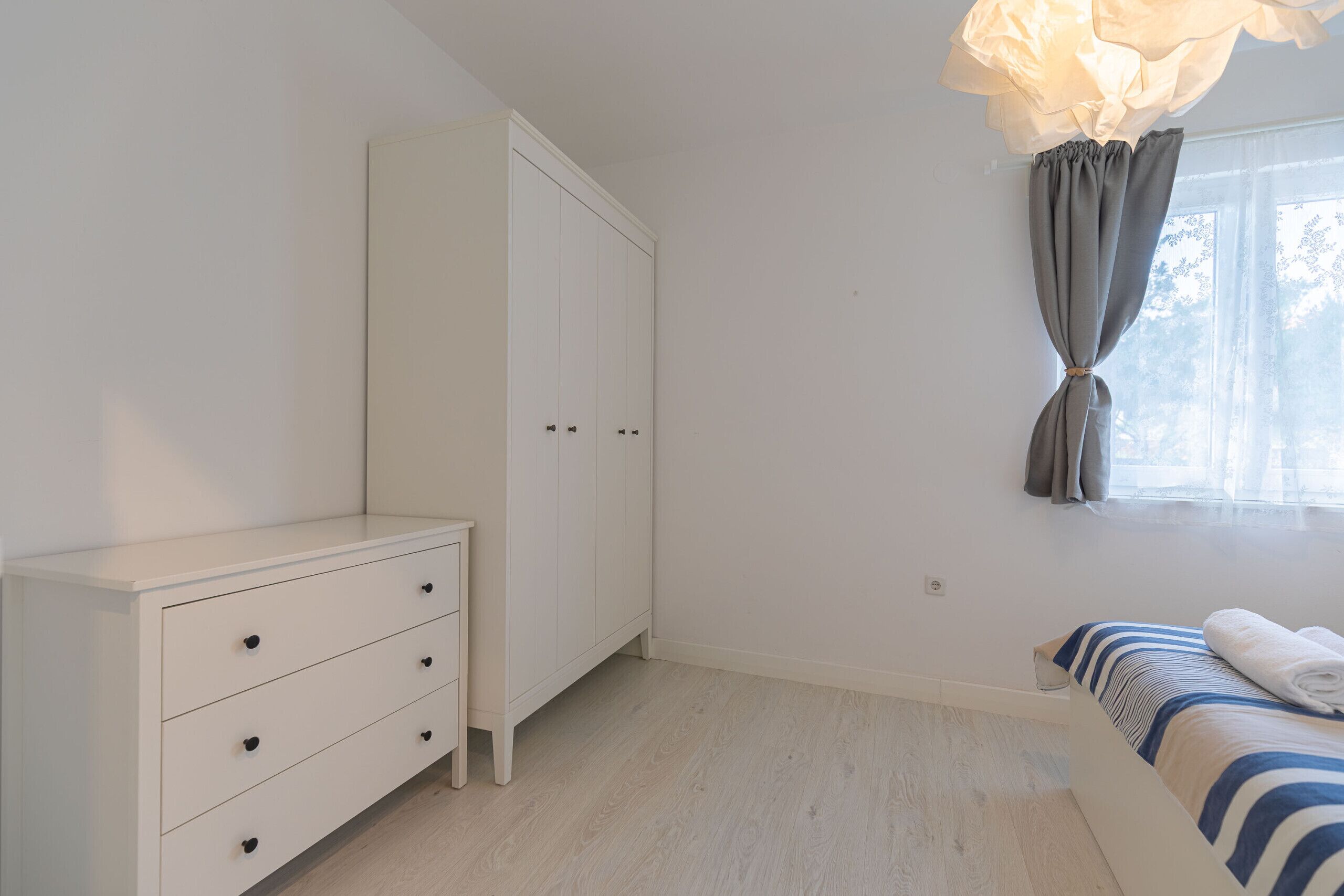 Une chambre avec une armoire blanche, un lit recouvert d’une courtepointe à rayures, une fenêtre avec des rideaux et un luminaire.