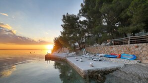 Ein Poolbereich am Wasser mit Liegestühlen, einer Steinmauer und Blick auf den Sonnenuntergang.