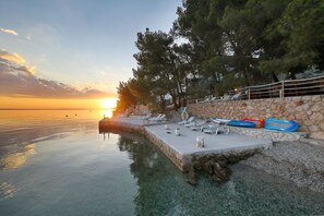 Ein Poolbereich am Wasser mit Liegestühlen, einer Steinmauer und Blick auf den Sonnenuntergang.