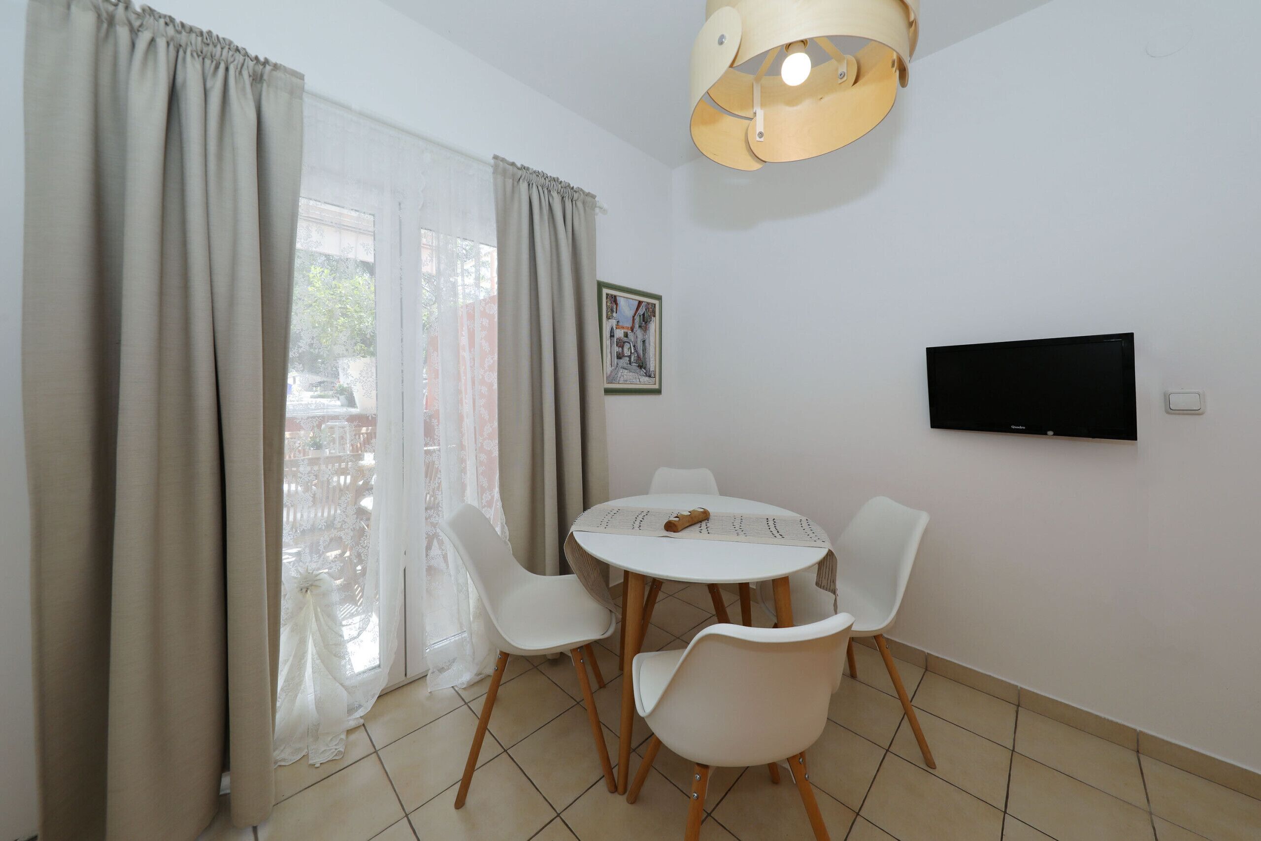 A room with a round table, four white chairs, a TV mounted on the wall, and a window with curtains.