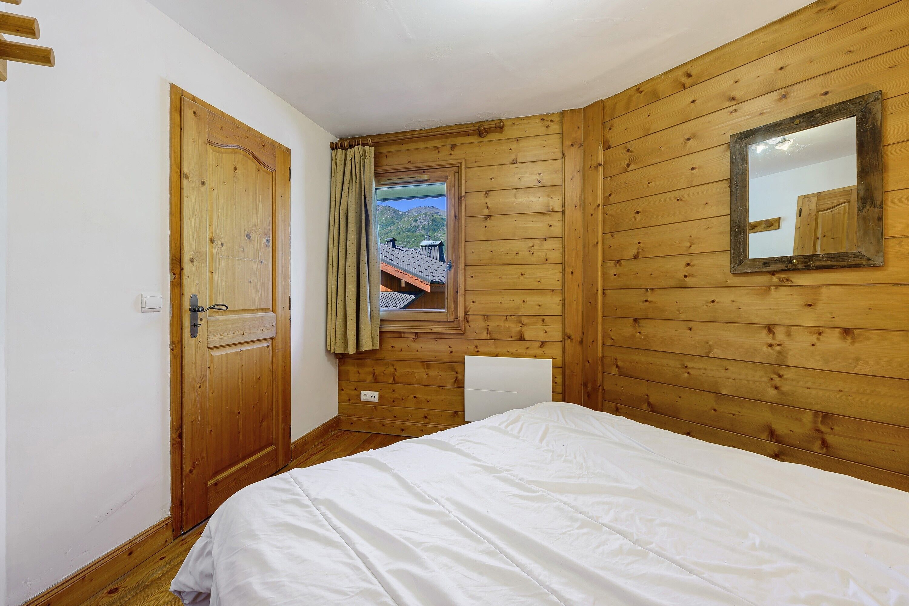 A bedroom with a wooden floor, a bed with white bedding, a wooden door, a window with a view of mountains, and a mirror on the wall.