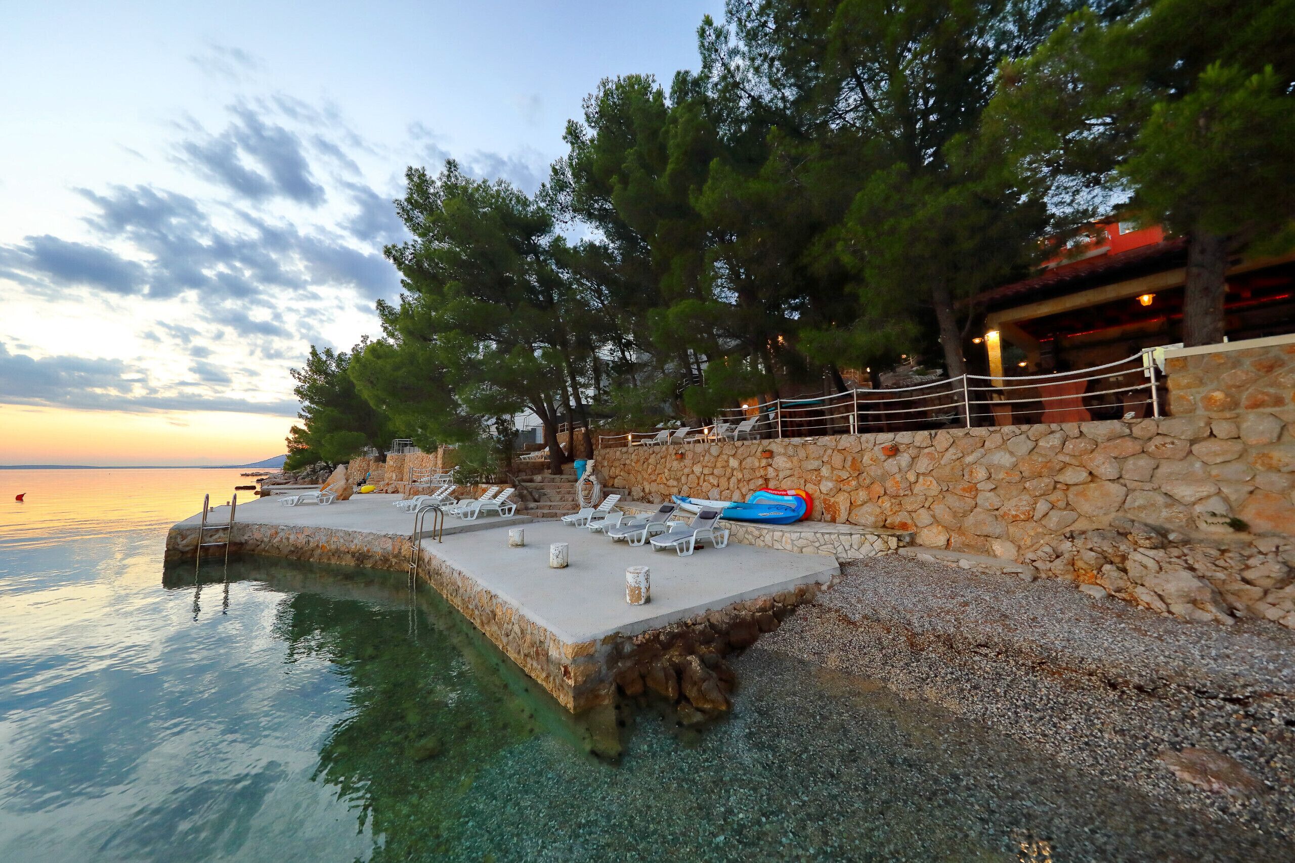 Un hôtel situé au bord de l’eau, doté d’un quai en pierre, de chaises longues et offrant une vue dégagée sur la mer.
