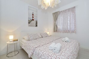 Ein Schlafzimmer mit Bett, Nachttisch, Lampe, einem Gemälde eines Leuchtturms und einem blumigen Vorhang.