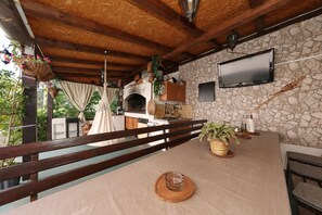 A rustic outdoor dining area with a stone wall, wooden beams, a television, and a brick oven. - Apartment Anita with Outdoor Kitchen (Zadar)