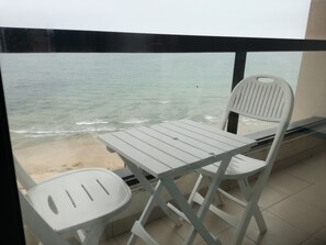 Ein Balkon mit weißem Tisch und zwei Stühlen, der Blick auf einen Strand mit ruhigem Wasser und eine Person in der Ferne gerichtet ist.
