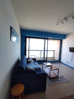 Een woonkamer met een bank, een bijzettafeltje, een stoel en een groot raam met balkon.