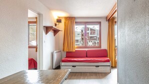 Quarto compacto com cama, uma mesinha, aquecedor e janela com cortinas.