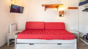 Uma cama com um edredom vermelho, uma mesinha de cabeceira, uma TV na parede e um abajur fixado na parede.