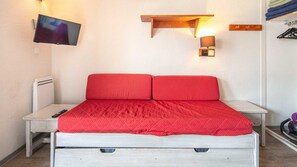 Ein Bett mit roter Decke, ein Nachttisch, ein an der Wand befestigter Fernseher und eine Wandlampe.