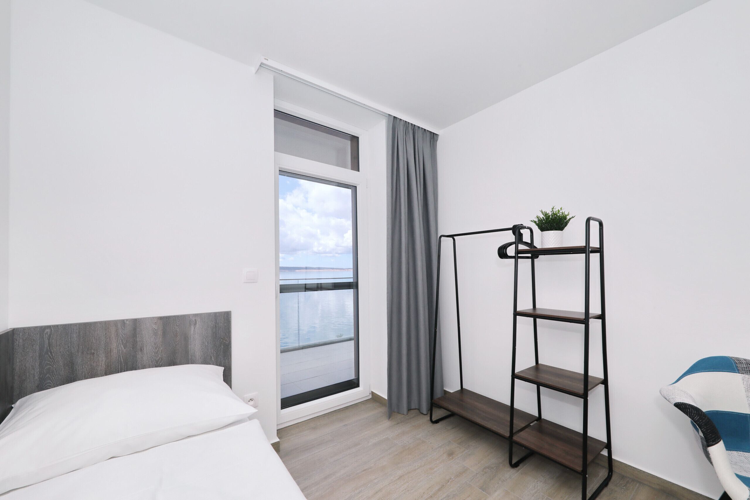 Un cuarto de hotel con cama, ventana con cortinas, una planta en un estante y vista al cielo.