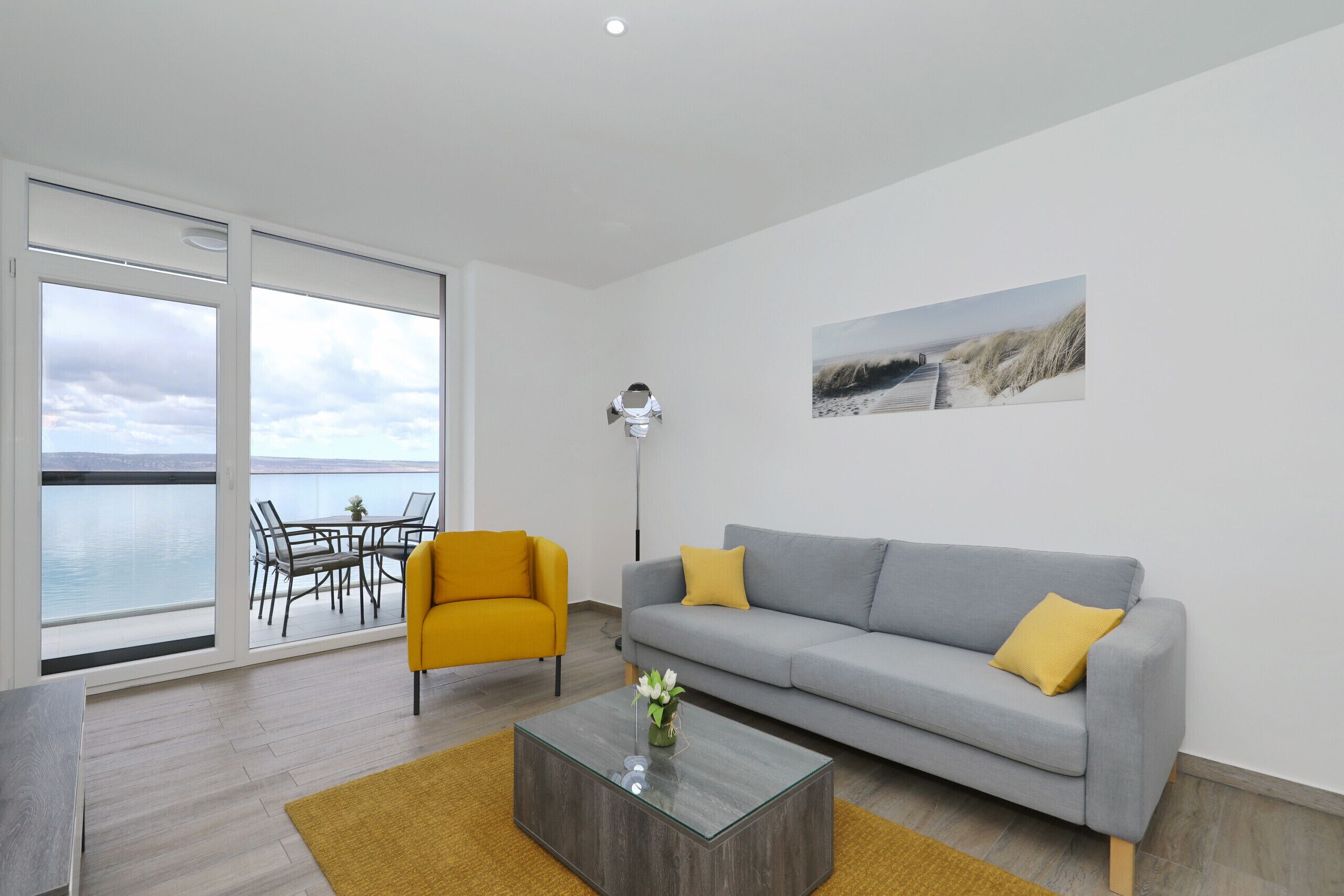 A modern living room with a sofa, a coffee table, a yellow armchair, and a view of the sea through large windows.