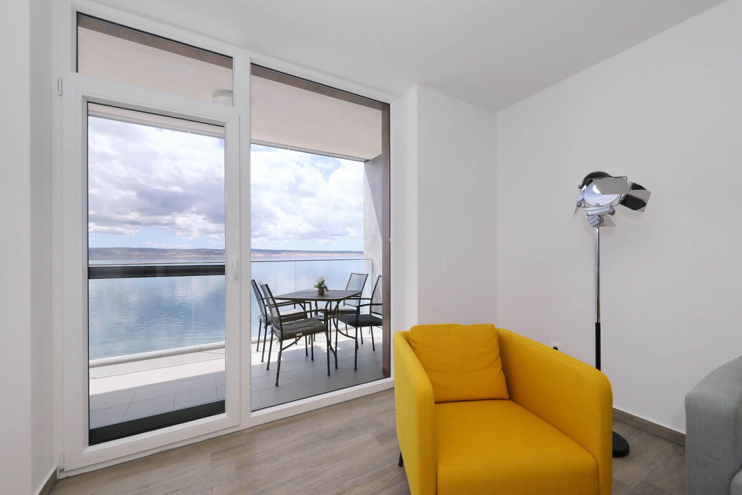 A room with a yellow sofa, a dining table with chairs, and a large window overlooking a body of water.