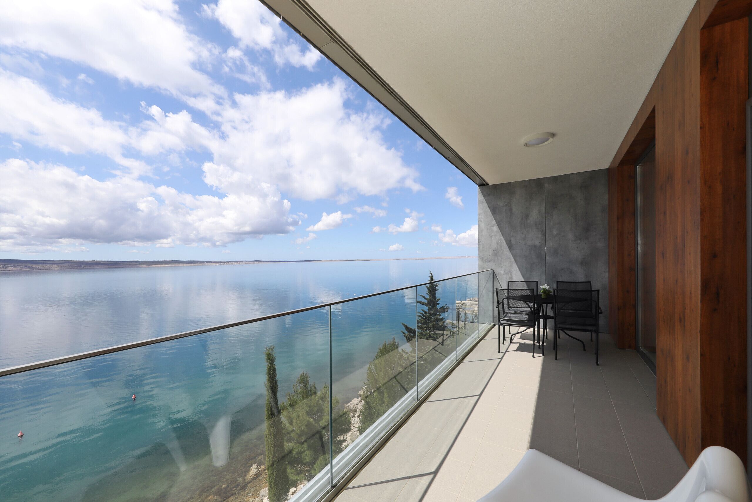 A balcony with a glass railing overlooking a body of water, a dining area with chairs and a table, and a wooden wall.