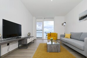 Ein modernes Wohnzimmer mit Flachbildfernseher, einer Couch, einem Couchtisch und Meerblick durch große Fenster.