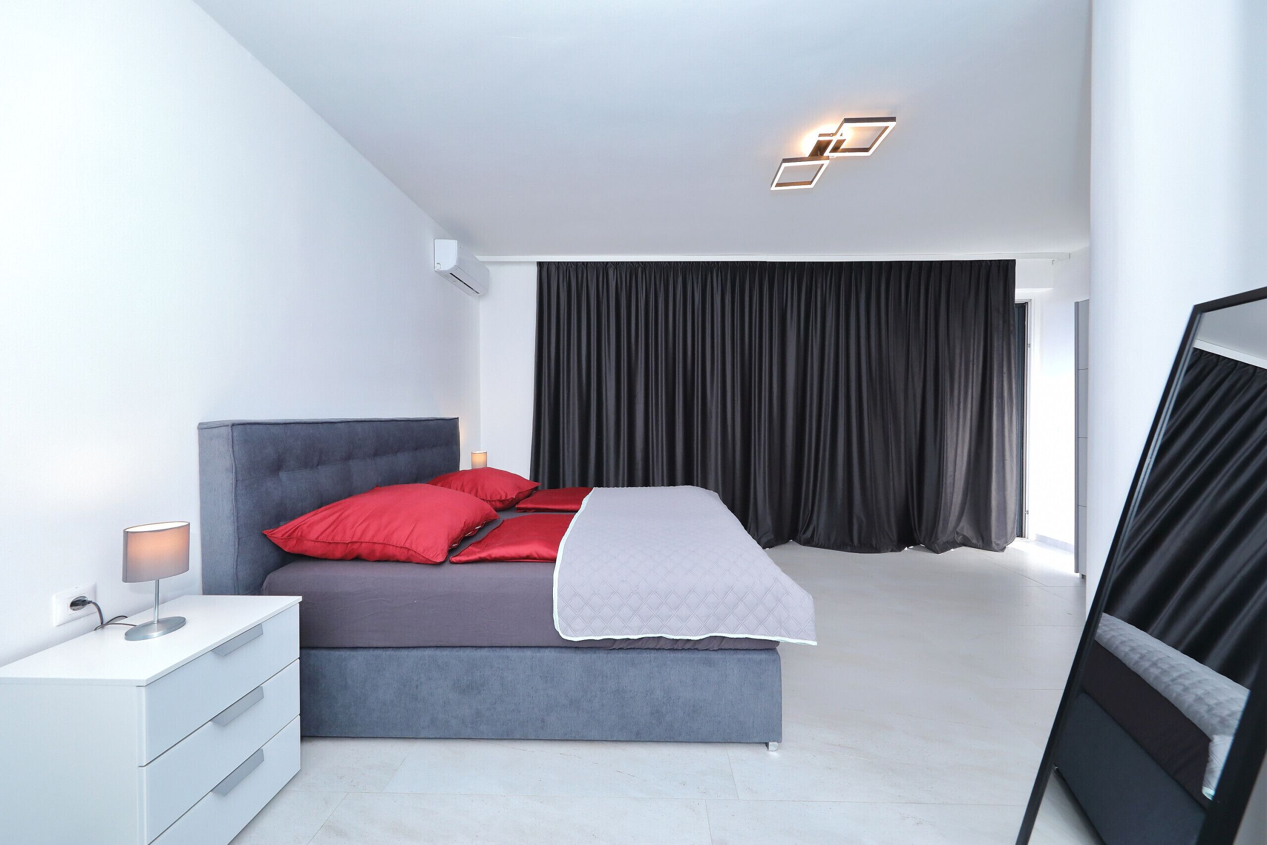 Un dormitorio moderno con una cama gris, almohadas rojas, una mesita de noche blanca y una cortina negra.