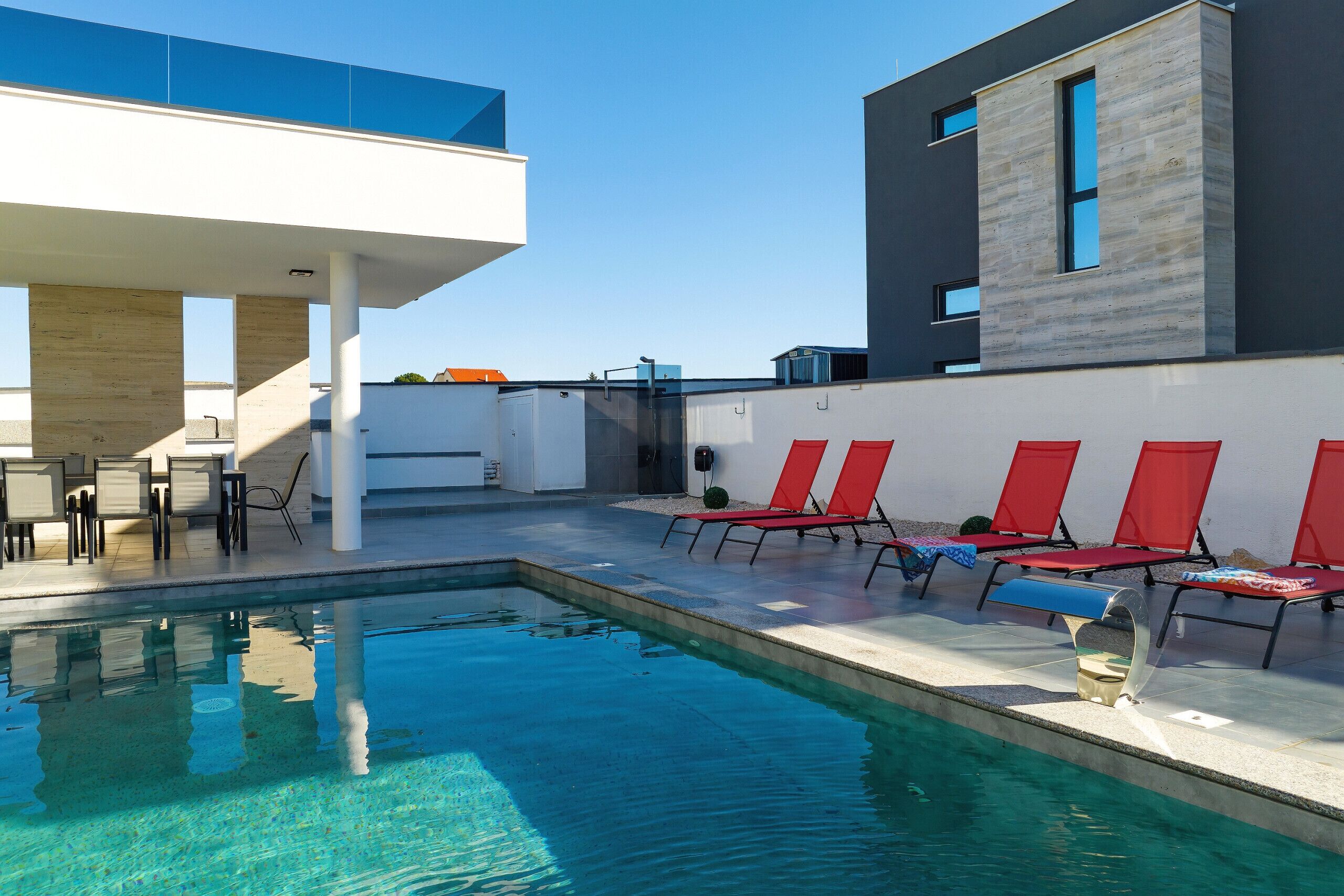 Une piscine bordée de chaises longues rouges, un espace de détente couvert et un bâtiment moderne doté de grandes fenêtres.