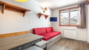 Una habitación con un banco de madera, un sofá rojo, un televisor montado y una ventana con vistas a montañas nevadas.