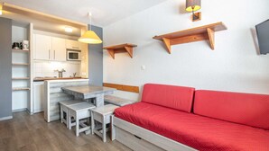 Un espacio habitable compacto con kitchenette, una mesa de comedor, un sofá rojo y un estante de madera pequeño.