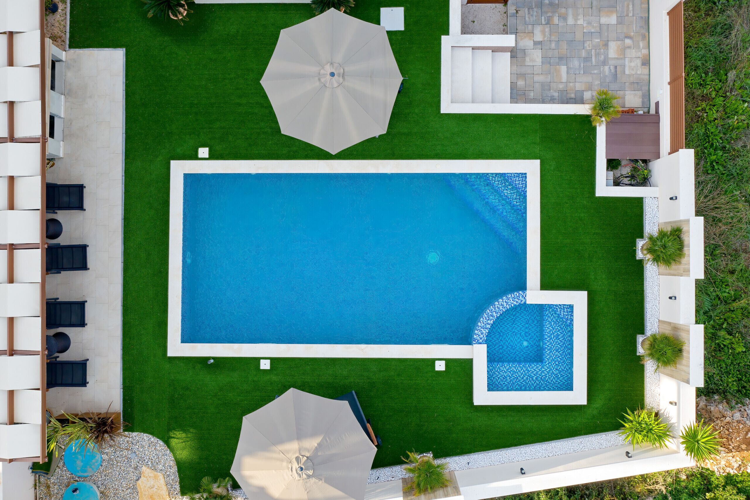 Ein rechteckiger Pool mit einem kleineren angrenzenden Pool, umgeben von einem gepflegten Rasen, Sitzmöglichkeiten im Freien und Sonnenschirmen.