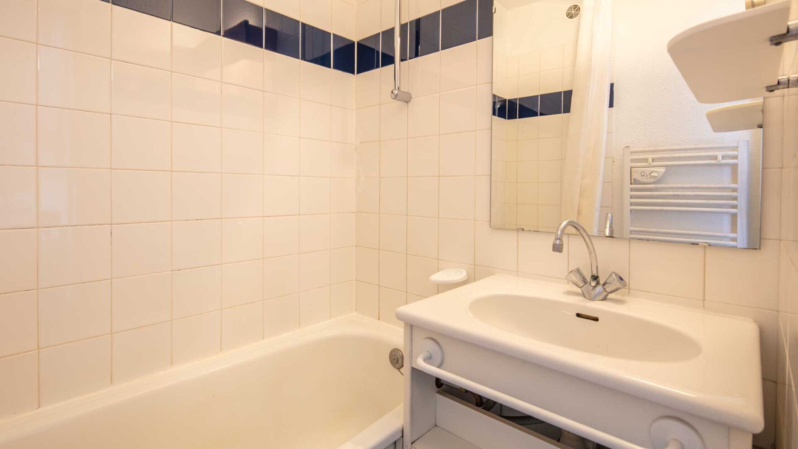 A bathroom with white tiles, a bathtub, a sink with a mirror, and a towel rack.