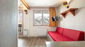 A room with a red sofa, a TV mounted on the wall, a window with a view of mountains, and a wooden shelf. - Charming studio for 4 people, with a balcony and a view of the ski slope (Tignes)