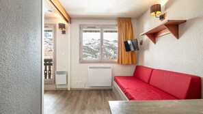A room with a red sofa, a TV mounted on the wall, a window with a view of mountains, and a wooden shelf. - Charming studio for 4 people, with a balcony and a view of the ski slope (Tignes)