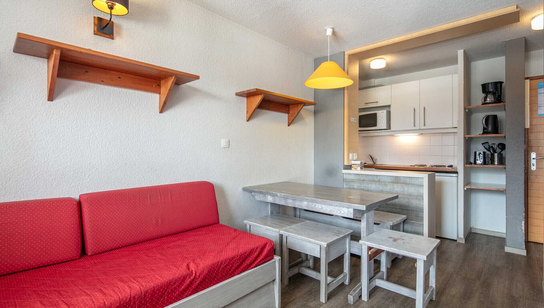A compact living space with a red sofa, a small dining table with benches, a kitchen area with white cabinets, and a yellow pendant light.