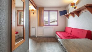 A room with a red sofa, a wooden table, a window with curtains, and a mounted TV. - Small, fully furnished studio for 4 people, with a balcony overlooking the ski slope (Tignes)