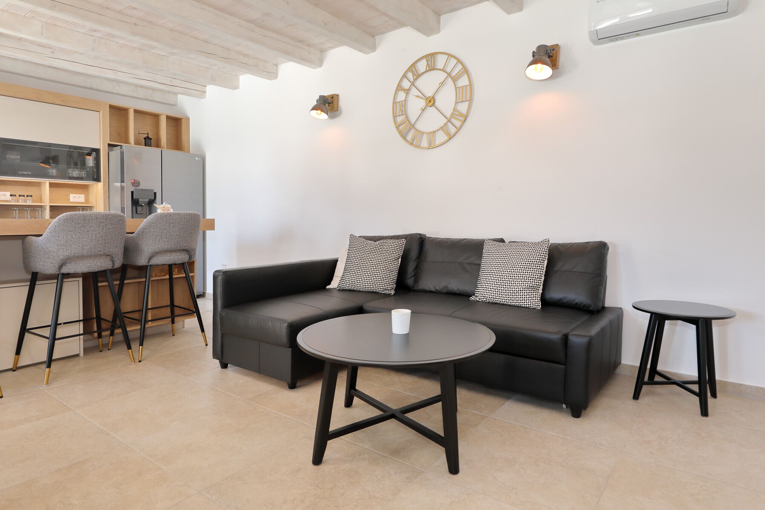 A modern living area with a sofa, two chairs, a coffee table, a wall clock, and a kitchen area with a refrigerator and a coffee machine.