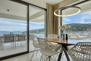 A modern dining area with a large table, white chairs, and a view of the sea and hills. - Apartment Mentis with Sea View (Marina)