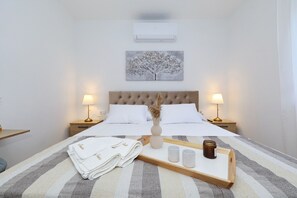 Ein ordentlich bezogenes Bett mit weißen Leinentüchern, ein Nachttisch mit Lampe, eine Wand-Klimaanlage und ein Bilderrahmen mit einem Baum.