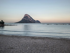 Uma praia com uma ilha rochosa ao longe, águas calmas e barcos.