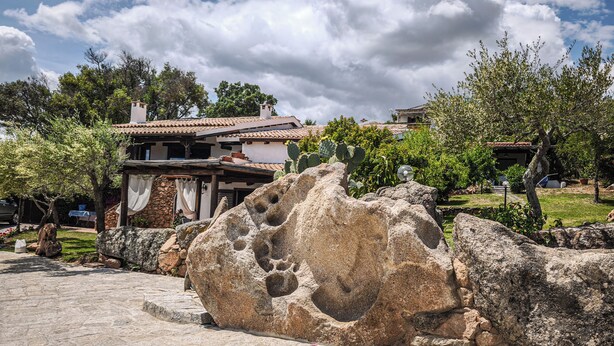 A stone pathway leading to a house with a tiled roof, surrounded by greenery and a large rock formation. - Monolocale in Villa (Porto San Paolo)