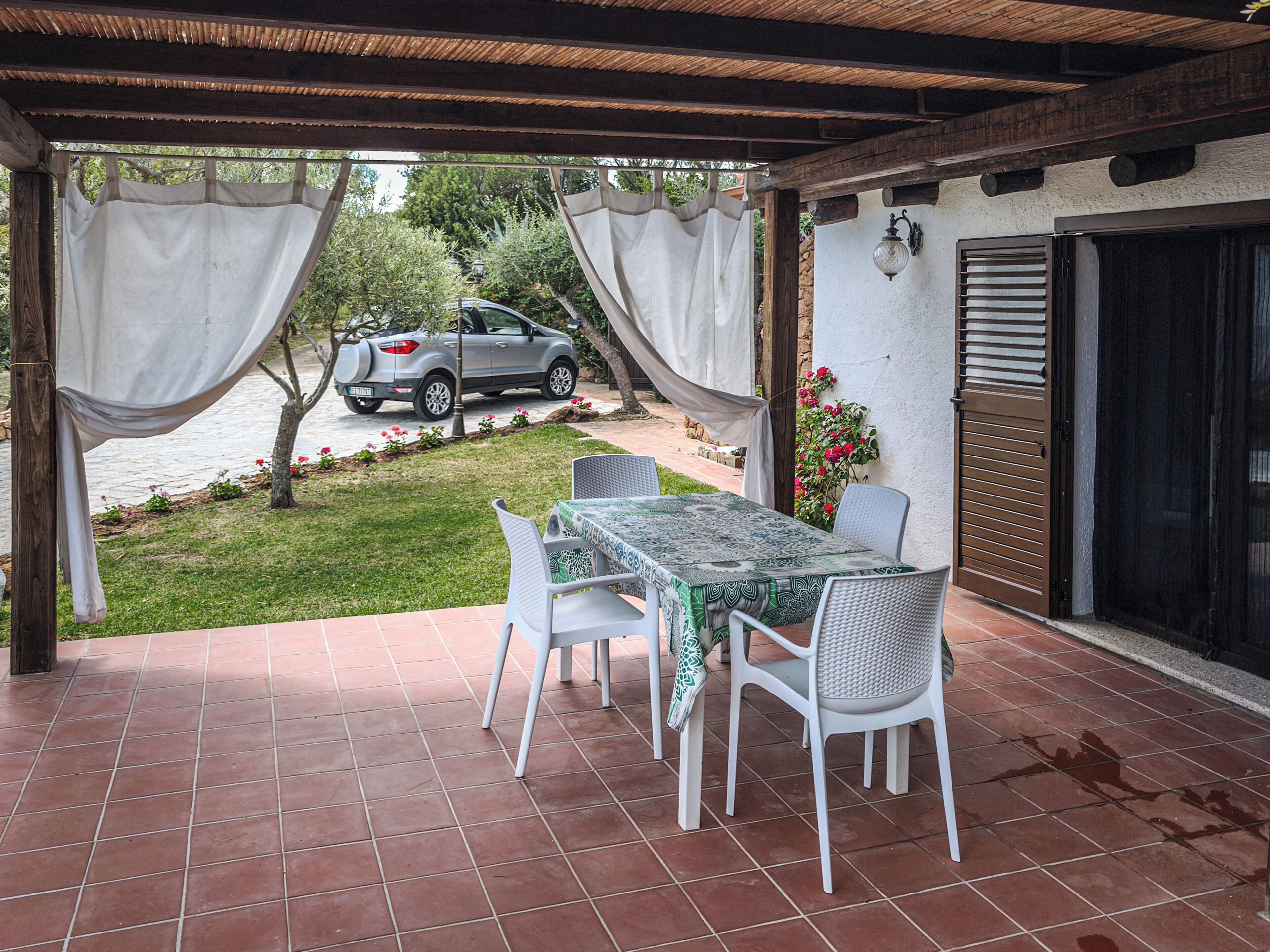 Un patio al aire libre con una mesa y sillas, un coche aparcado al fondo y vistas a un jardín.
