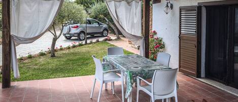 Un patio al aire libre con una mesa y sillas, un coche aparcado al fondo y vistas a un jardín.