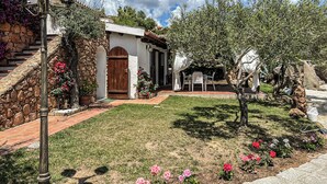 A stone building with a wooden door, a small garden with flowers, and a statue. - Monolocale in Villa (Porto San Paolo)