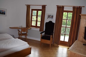 A room with a bed, a wooden chair, a table with a glass, and a chair with a black cushion.