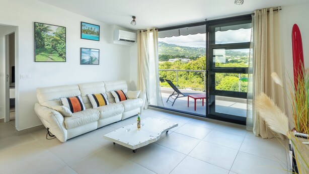 A living room with a white sofa, a coffee table, and a large sliding glass door leading to a balcony with outdoor furniture. - Atea Pearl (Pirae)