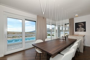 A modern dining area with a large wooden table, white chairs, a view of a pool, and a mountain landscape.