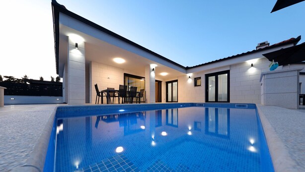Una piscina con solárium alicatado, un porche cubierto con sillas y una puerta que da a un espacio interior.