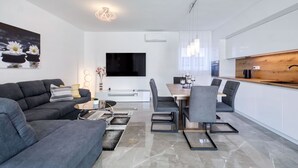 Una sala de estar moderna con sofá, mesa de centro y televisor. Hay un comedor con mesa y sillas. La cocina tiene una encimera de madera y armarios blancos.