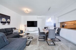 A modern living room with a sofa, coffee table, and TV. There's a dining area with a table and chairs. The kitchen has a wooden countertop and white cabinets. - Villa Mariva with Heated Pool (Vir)