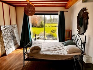 A bedroom with a bed, a hanging light fixture, a view of a garden, and a decorative wall piece.