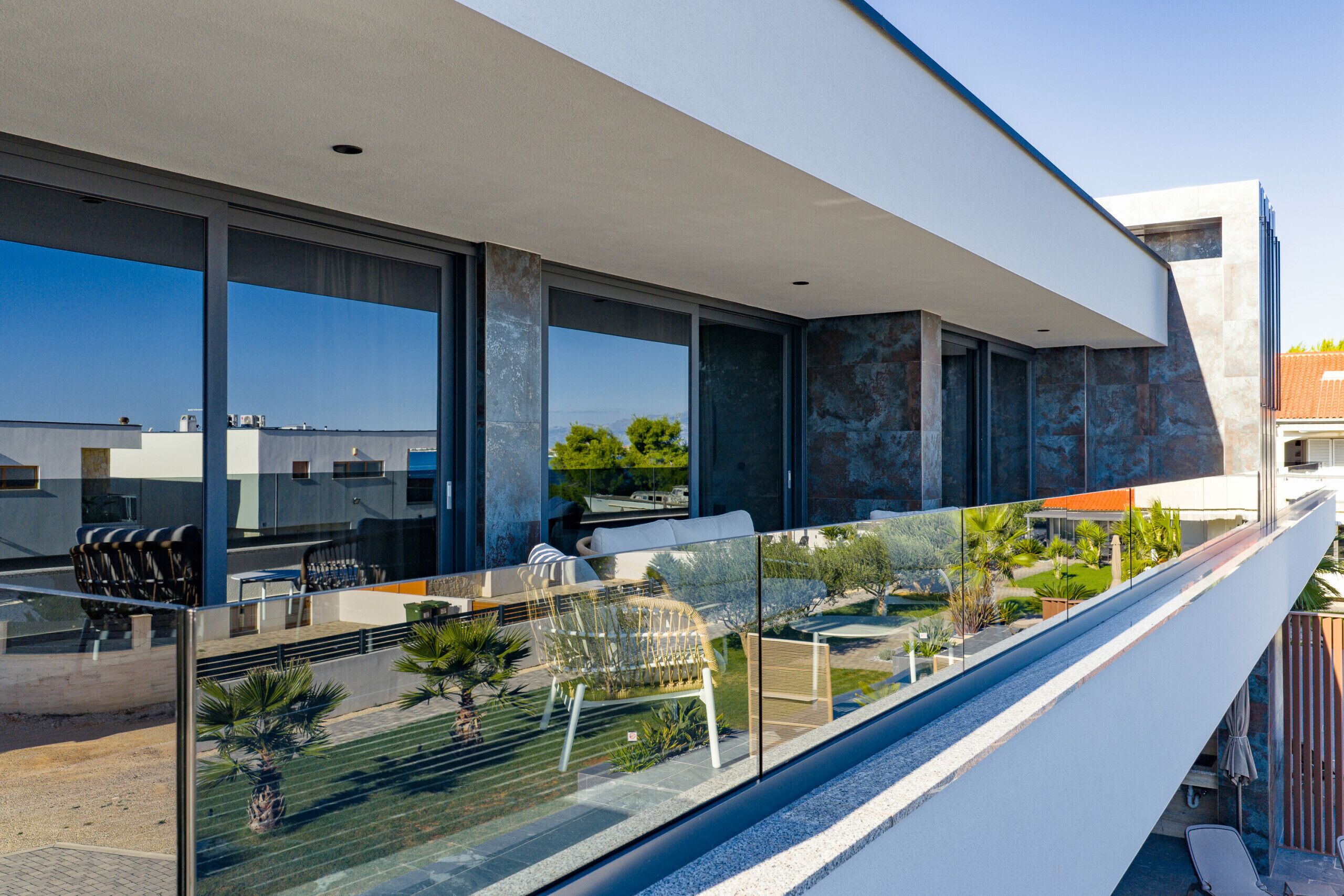A modern building with large windows, a balcony with seating, and a view of a landscaped garden.