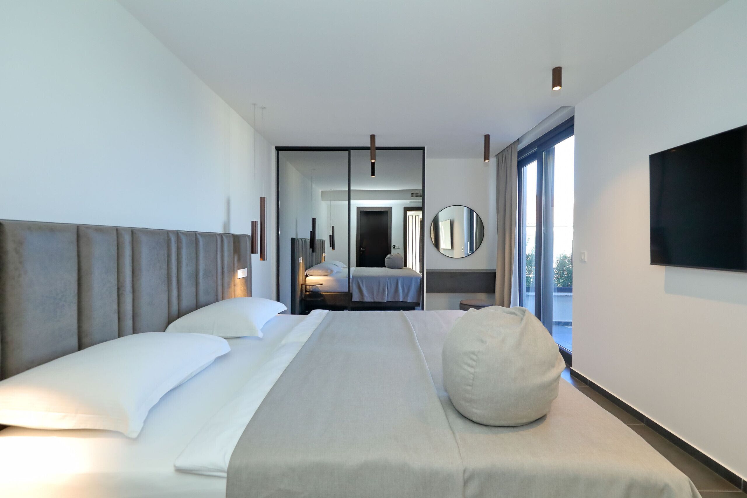 Quarto de hotel moderno com uma cama grande, um espelho redondo, uma televisão de tela plana e vista para o exterior.
