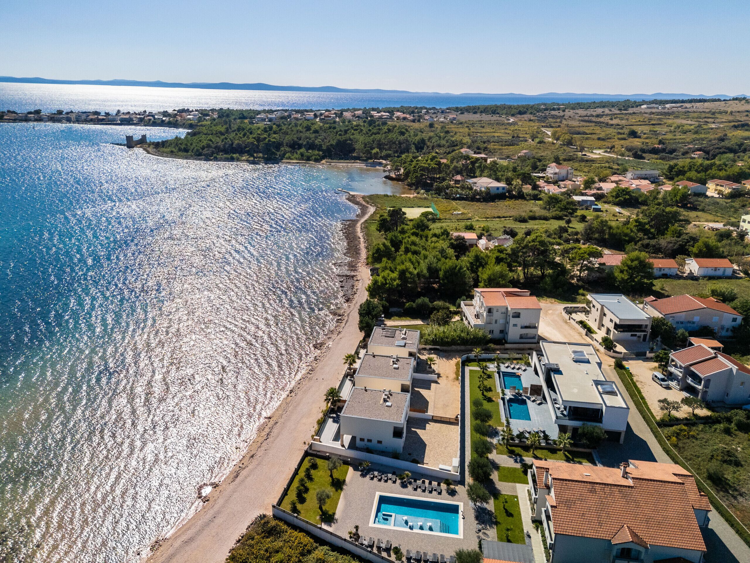 An aerial view of a coastal area with a beach, a swimming pool, and residential buildings.