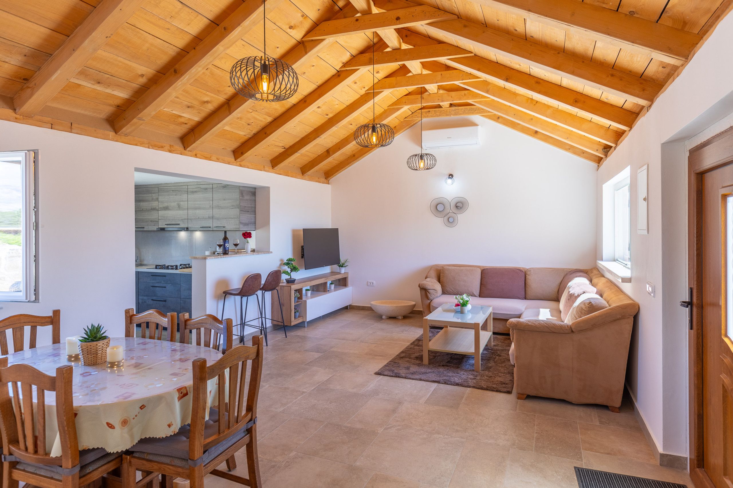 Amplia sala de estar con techo alto de madera, una mesa de comedor con sillas, un sofá y una cocina al fondo.